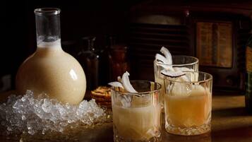 A bottle and two glasses of a beverage with ice and a garnish, placed on a table with a dark background and vintage decor.