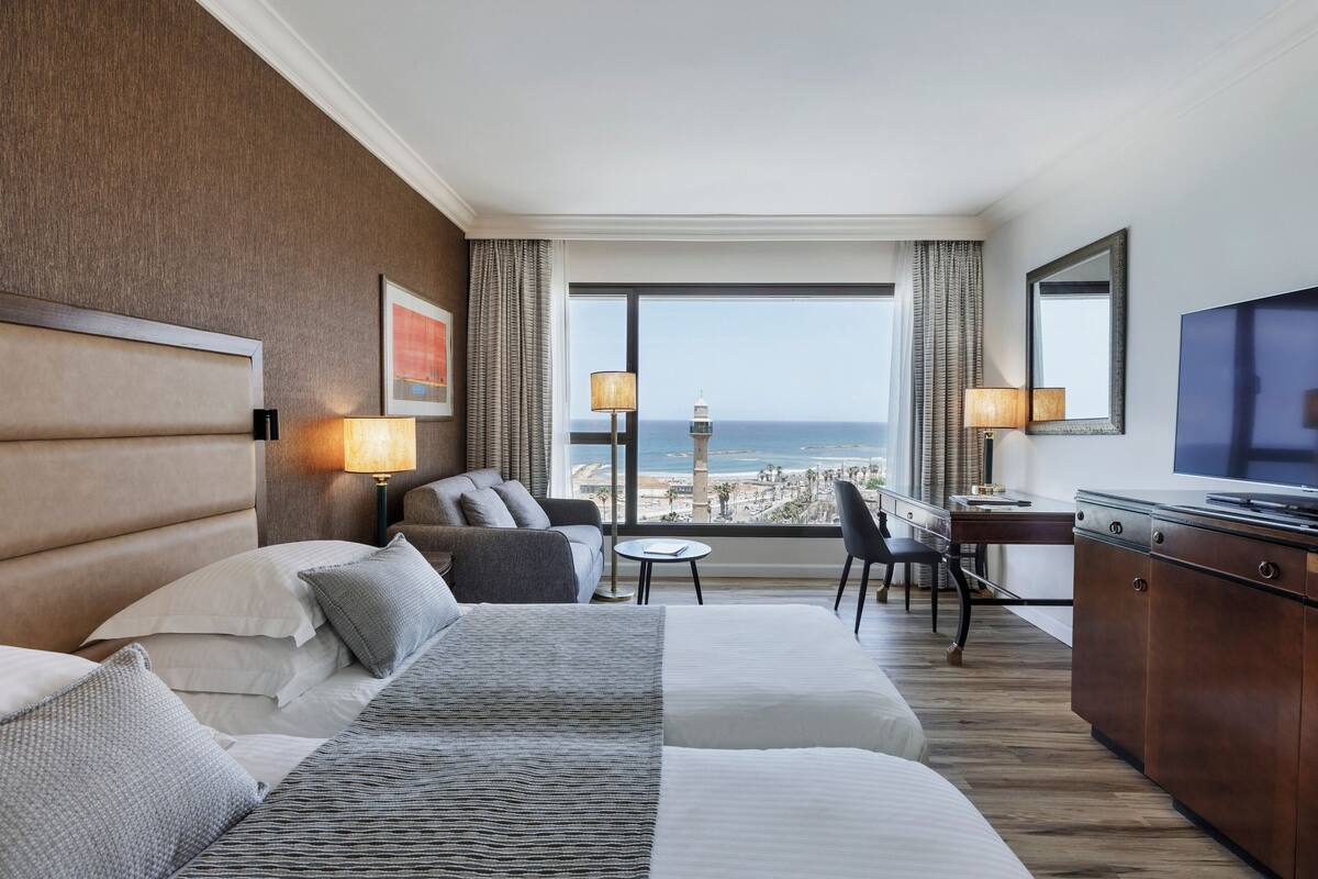 Habitación de hotel con una cama grande, un escritorio, una silla y un televisor. Se ve el océano a través de la ventana.