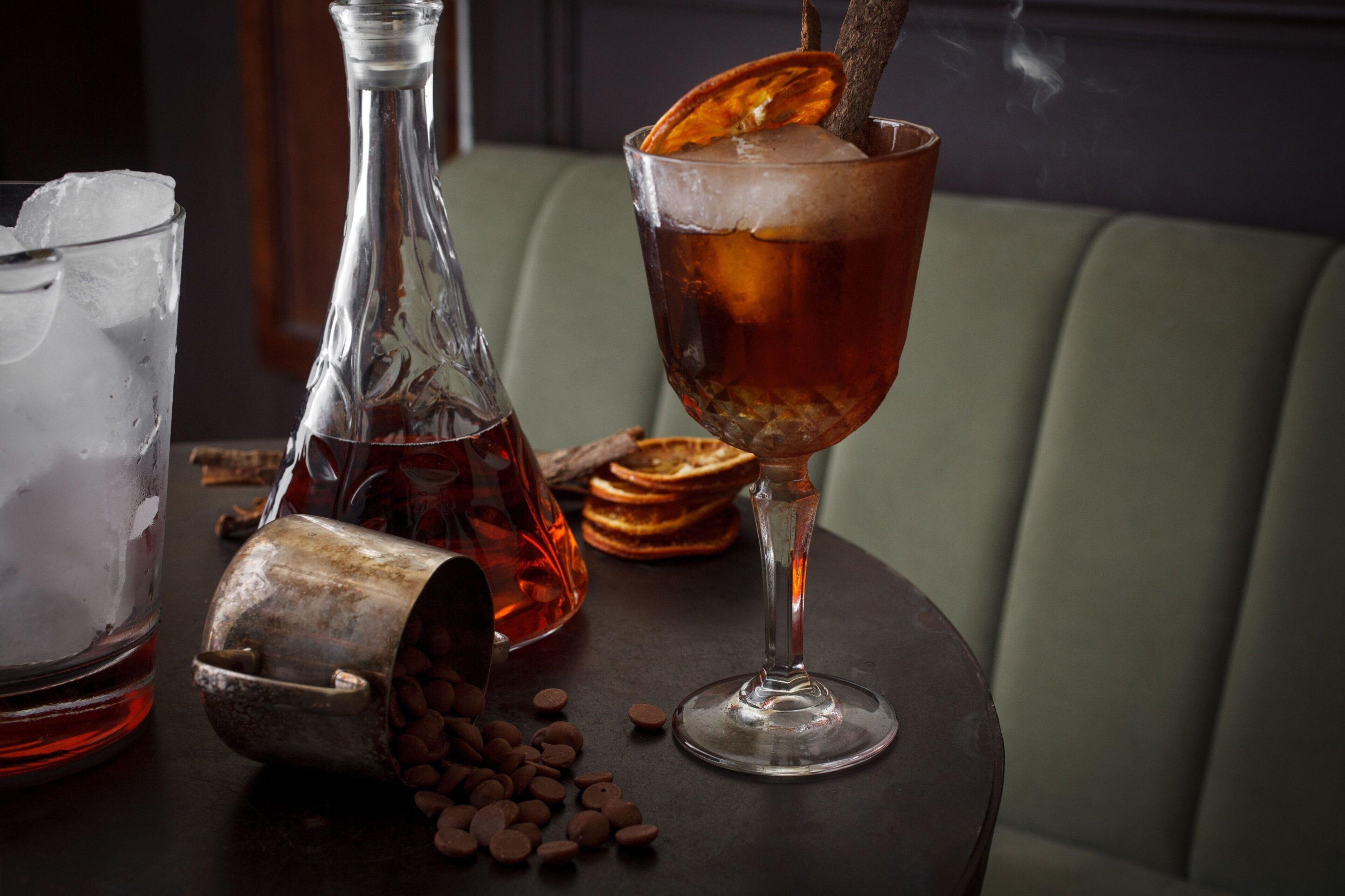 Un vaso de bebida color ámbar con hielo y una rodaja de cáscara de naranja, una botella de líquido rojo y trozos de chocolate esparcidos sobre una mesa.