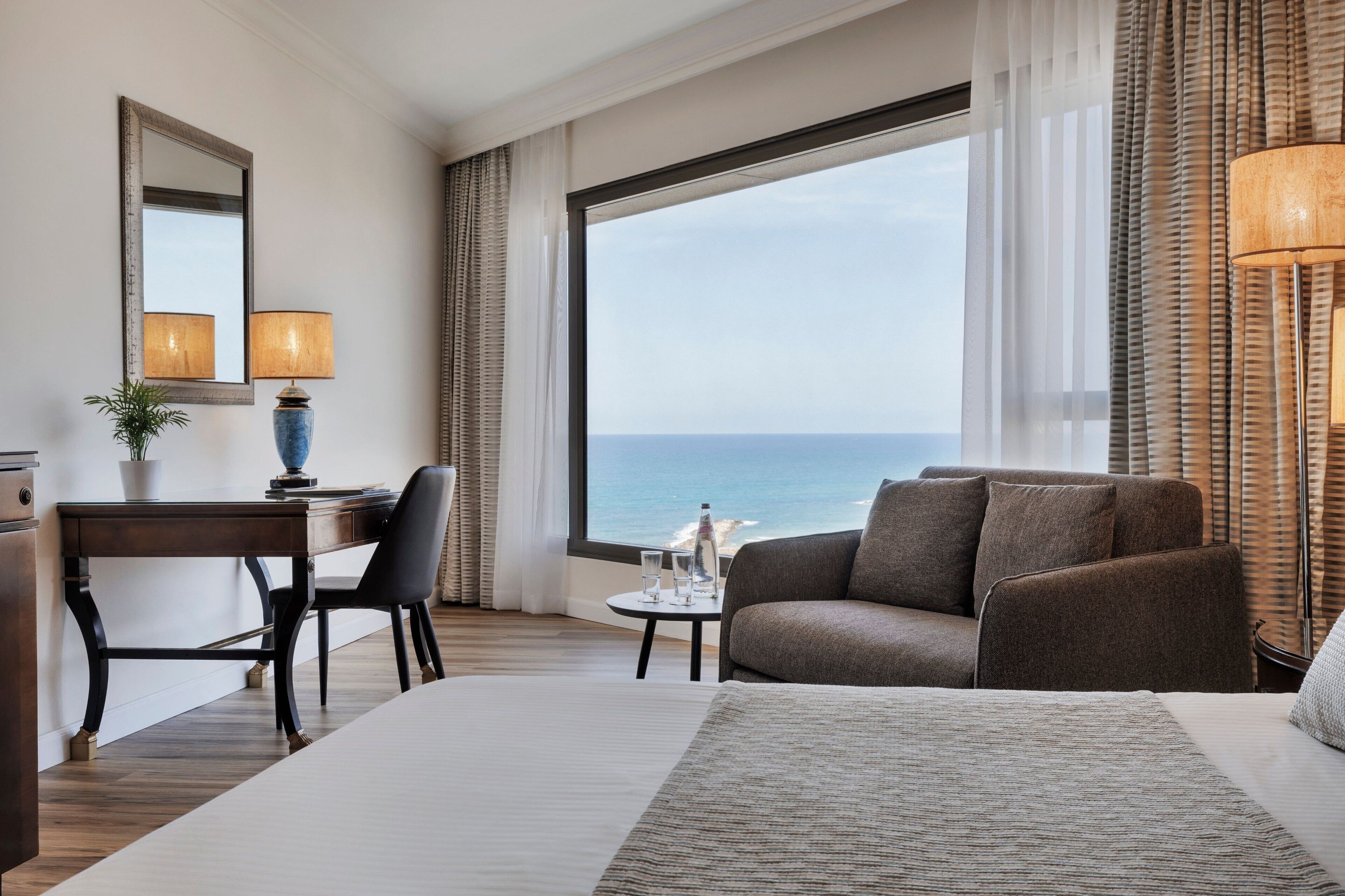 A hotel room with a large bed, a desk with a chair, a small table with a lamp, and a view of the sea through large windows.