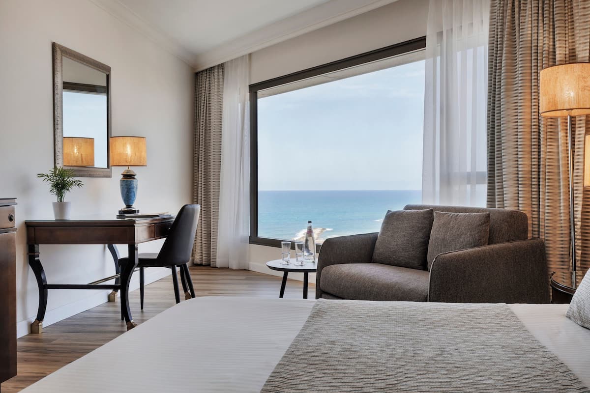 Habitación de hotel con una cama grande, un escritorio con silla, una mesita con lámpara y vista al mar a través de amplias ventanas.