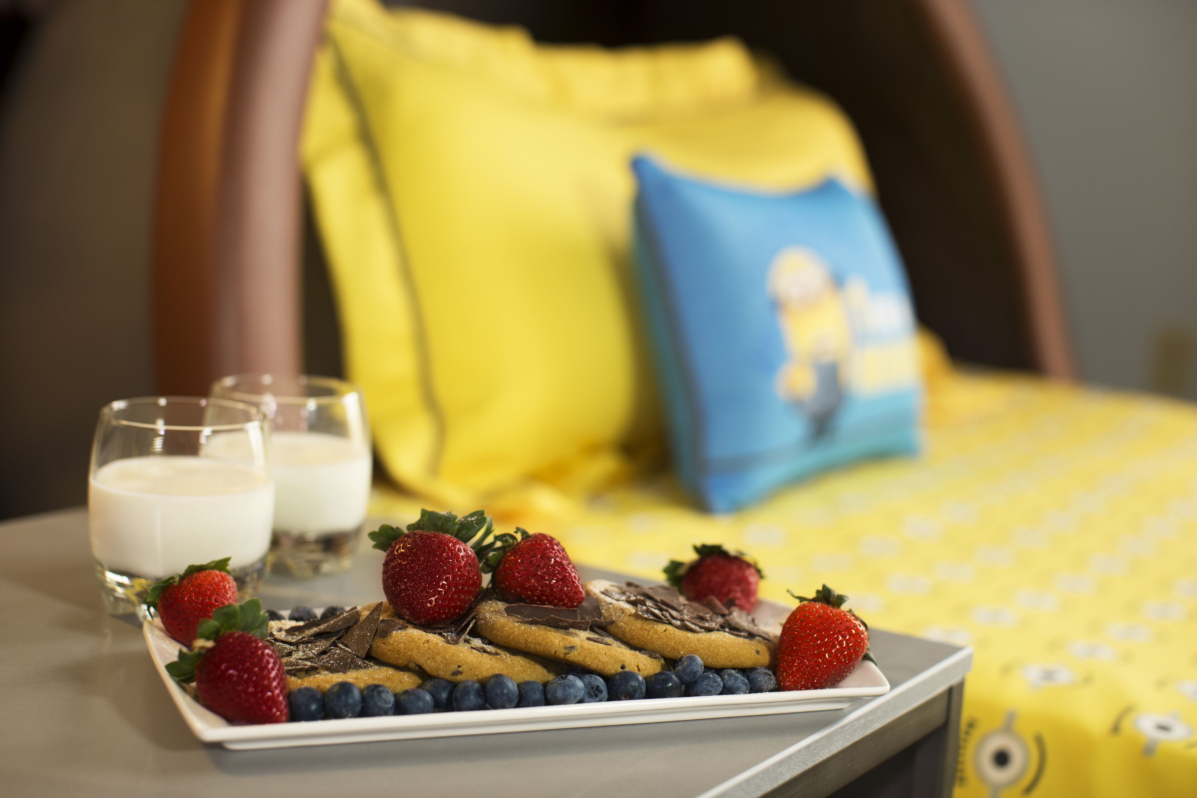 A breakfast tray with cookies, strawberries, and blueberries on a white plate, accompanied by two glasses of milk, set on a table in a room with yellow bedding and a blue pillow.