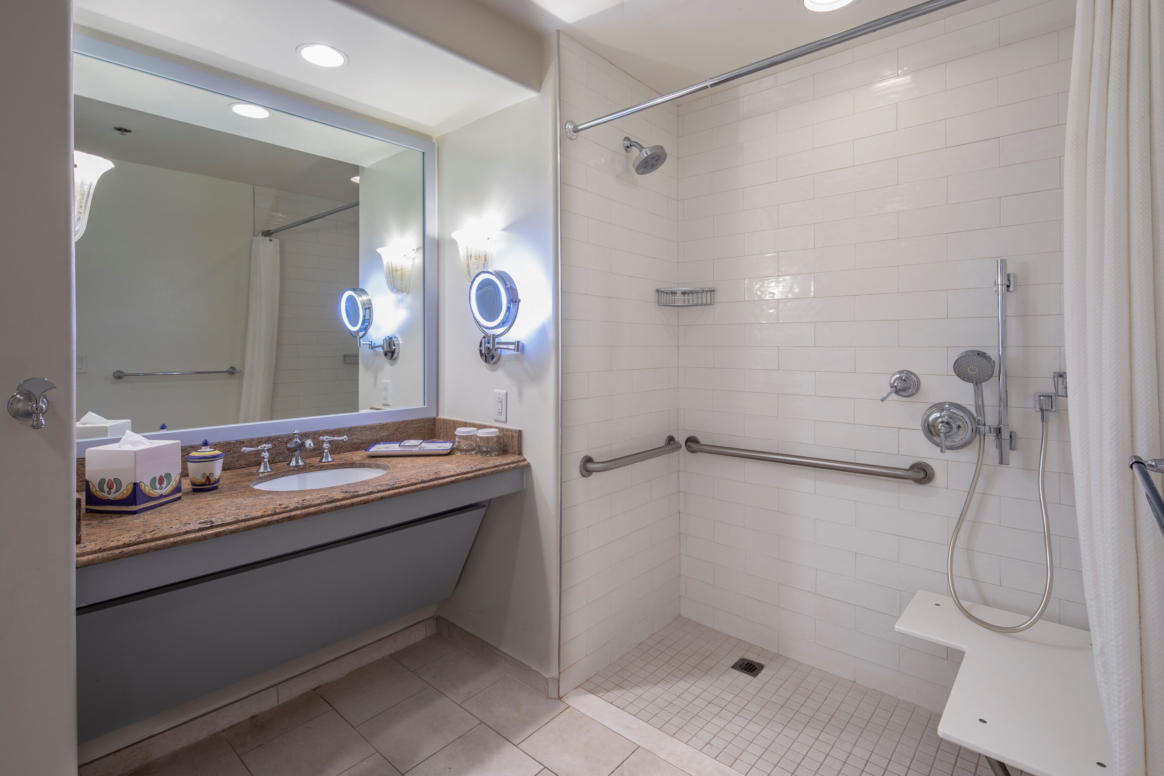 Un baño moderno con un espejo grande, dos lavamanos, ducha con barras de apoyo y un inodoro.