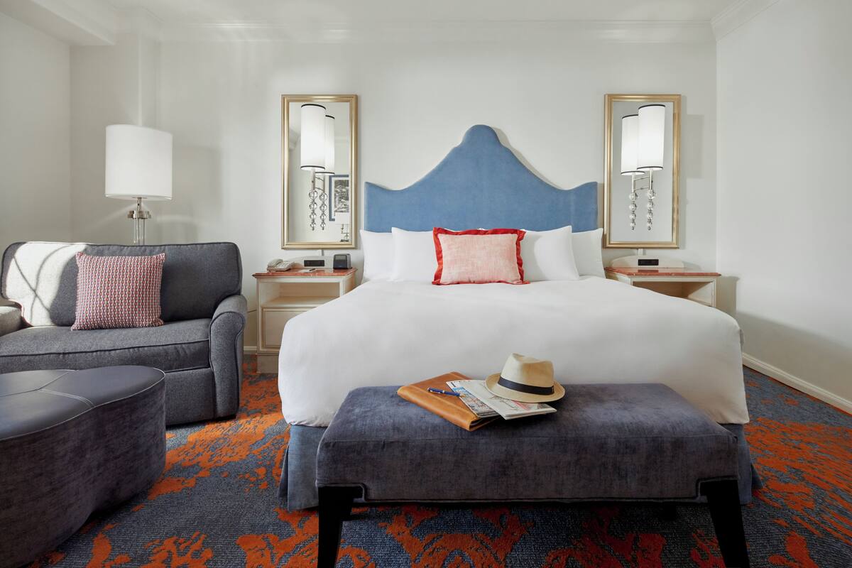 Une chambre d’hôtel avec un grand lit, un canapé gris, un pouf assorti, une table de chevet avec une lampe et deux miroirs fixés au mur.