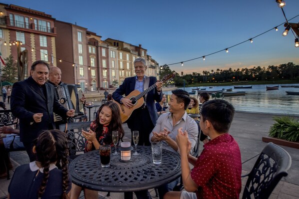 En grupp människor sitter runt ett bord utomhus, och en musiker spelar gitarr.