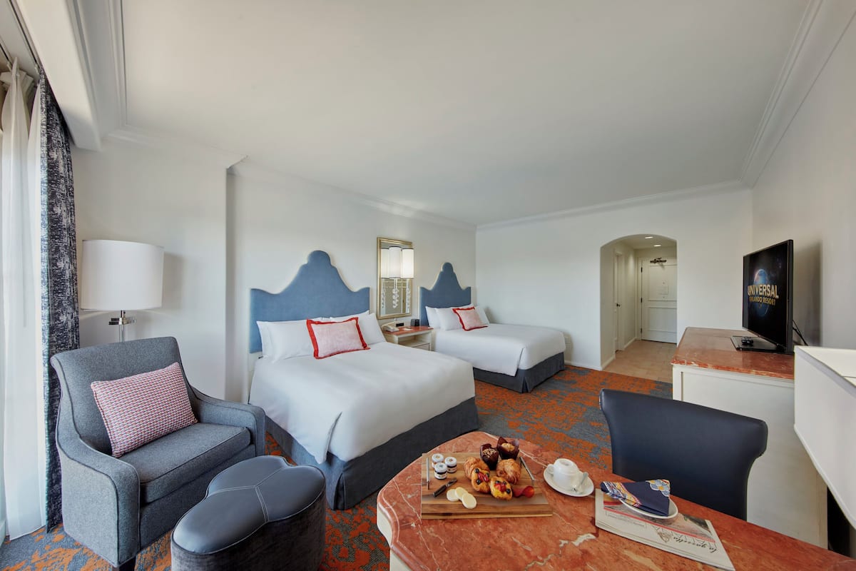 Ein Hotelzimmer mit zwei Betten, einem Schreibtisch, einem Sessel, einem Fernseher und einem Esstisch mit Speisen.