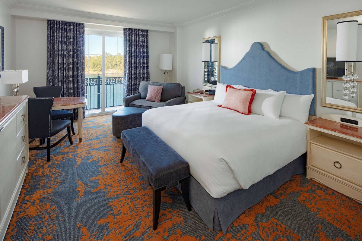 Une chambre d’hôtel avec un grand lit, un bureau avec une chaise, un banc, une table de chevet et une fenêtre avec des rideaux.