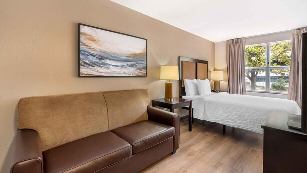 Ein Hotelzimmer mit braunem Ledersofa, einem Bett mit weißer Bettwäsche, einem Nachttisch mit Lampen und einem großen Fenster mit Vorhängen.