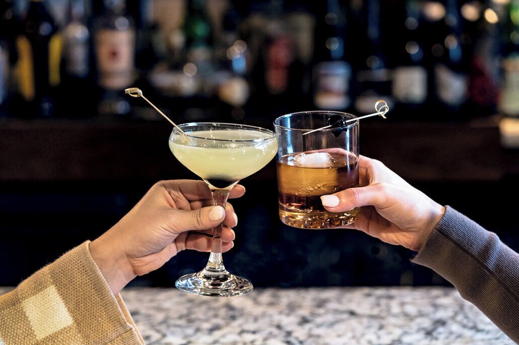 Dos manos sosteniendo copas de cóctel con pajitas, una con martini y la otra con whiskey, en un bar con botellas al fondo.