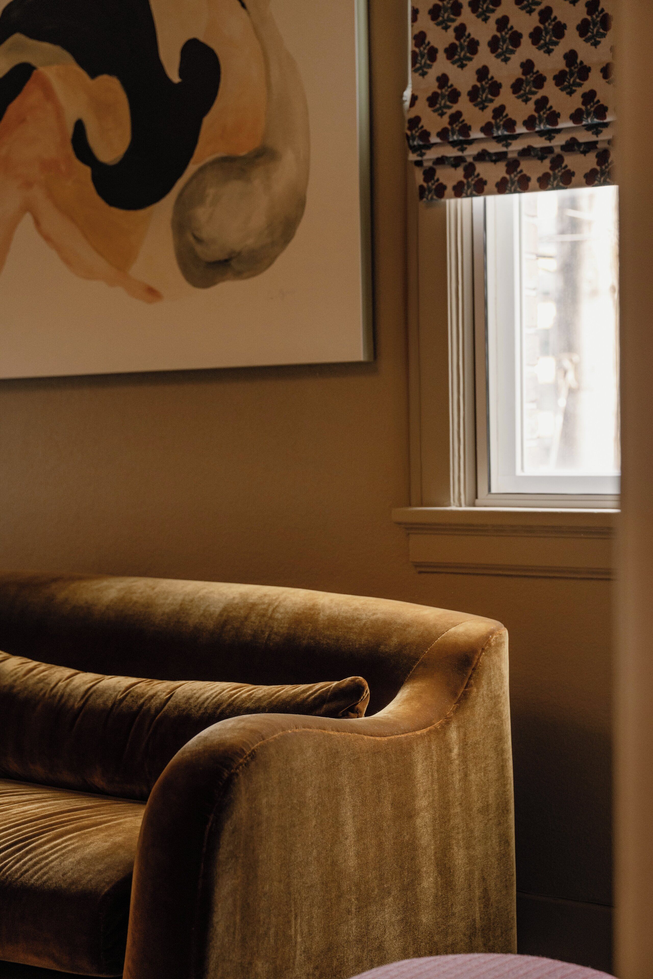 A room with a brown sofa, a window with a patterned valance, and a framed artwork on the wall.