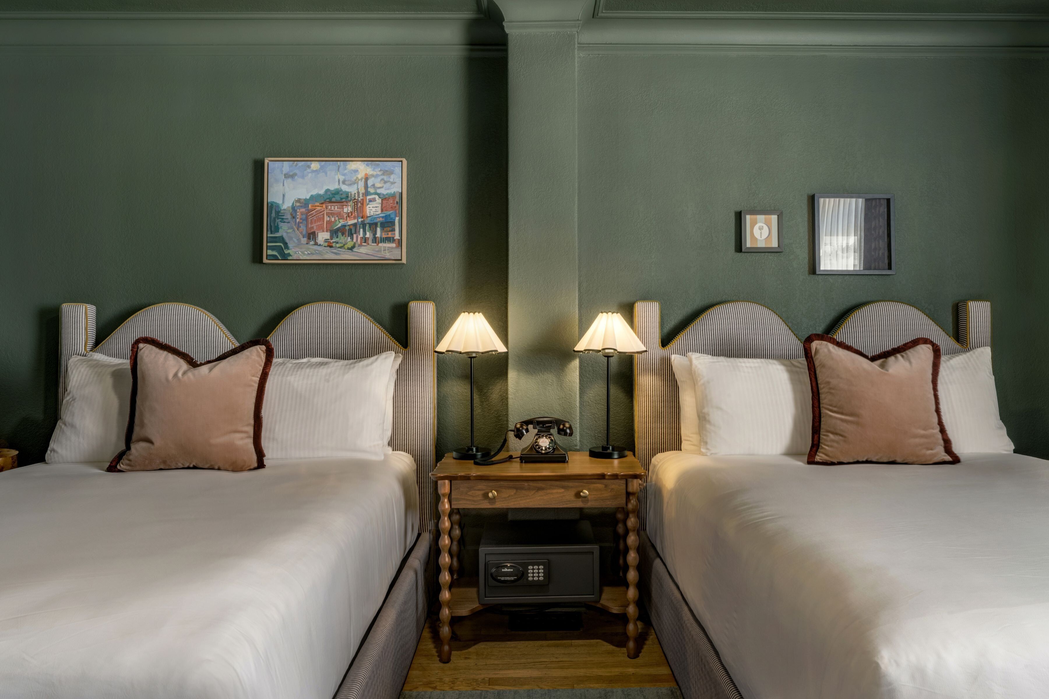 Habitación de hotel con dos camas, una mesita de noche con teléfono y caja fuerte, y obras de arte en las paredes.