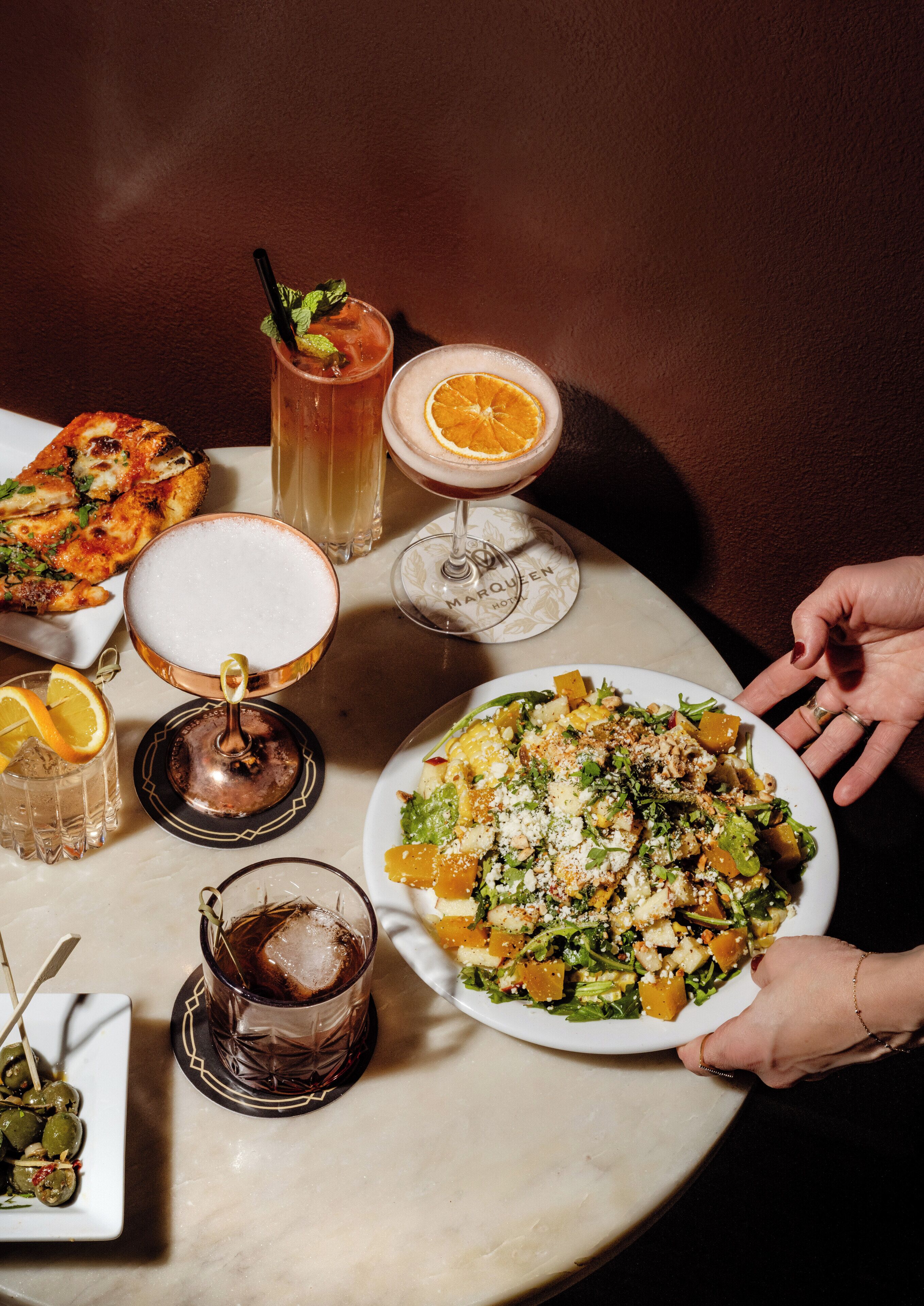 A table with various dishes and drinks, including a plate of salad, a glass of iced tea, and a cocktail.