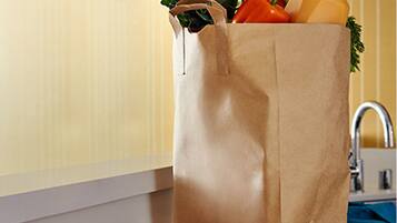 A brown paper grocery bag filled with vegetables and a bottle of juice on a kitchen counter.
