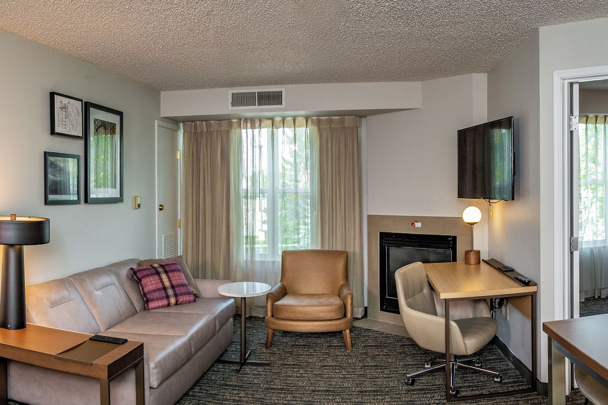 A hotel room with a bed, a sofa, a desk, a chair, a lamp, a fireplace, and a television.