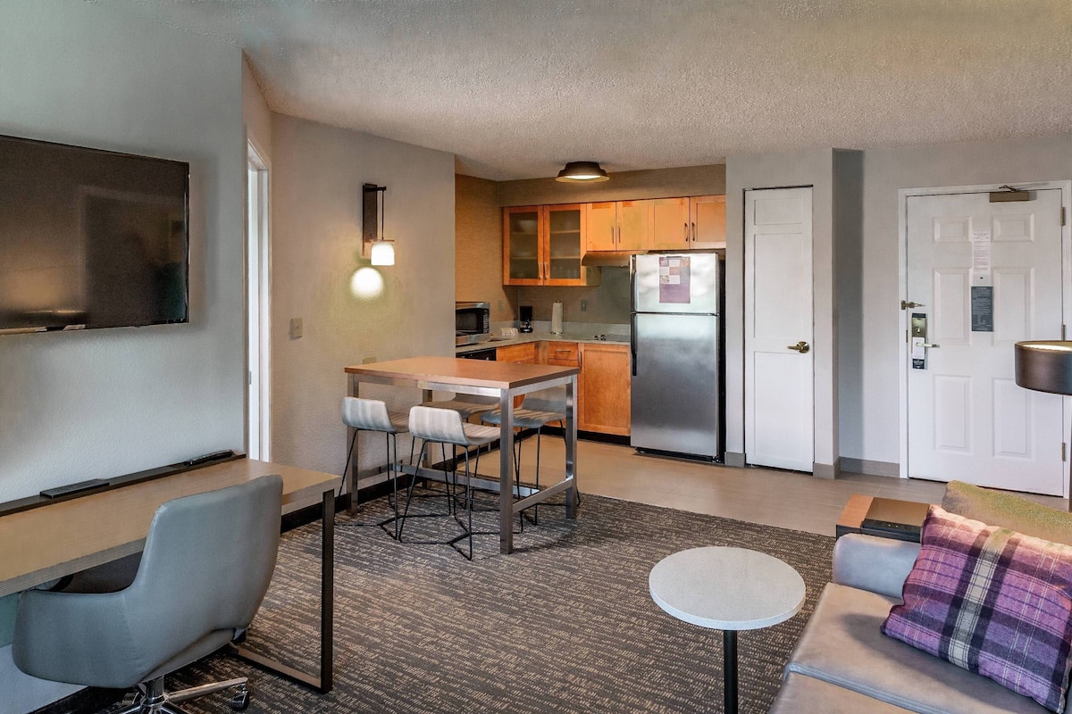 A compact living space with a kitchenette, a dining area with bar stools, a sofa with a plaid throw, and a flat-screen TV mounted on the wall.