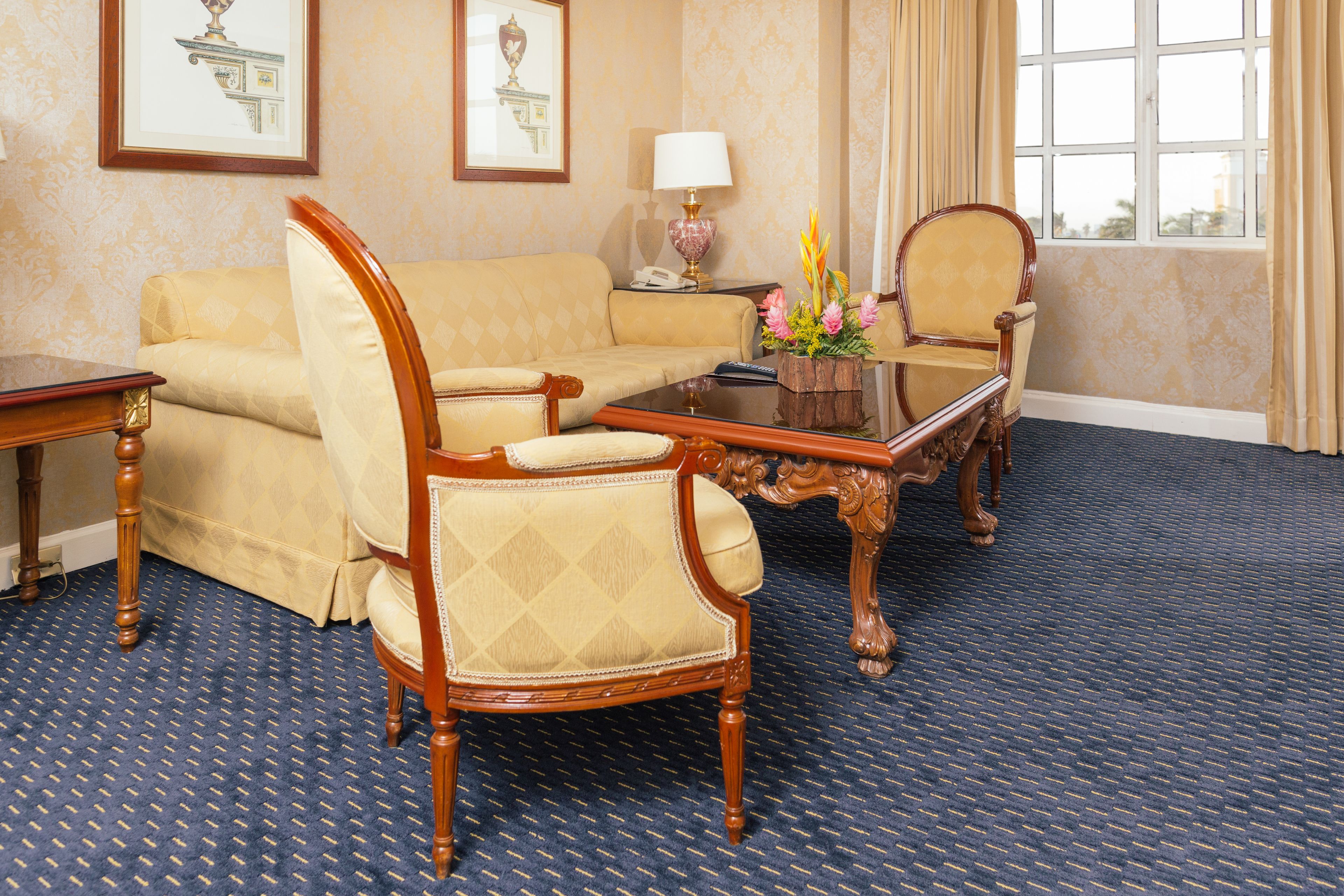 Una habitación con alfombra azul, un sofá color beige, una mesa de centro de madera y dos sillas de madera.