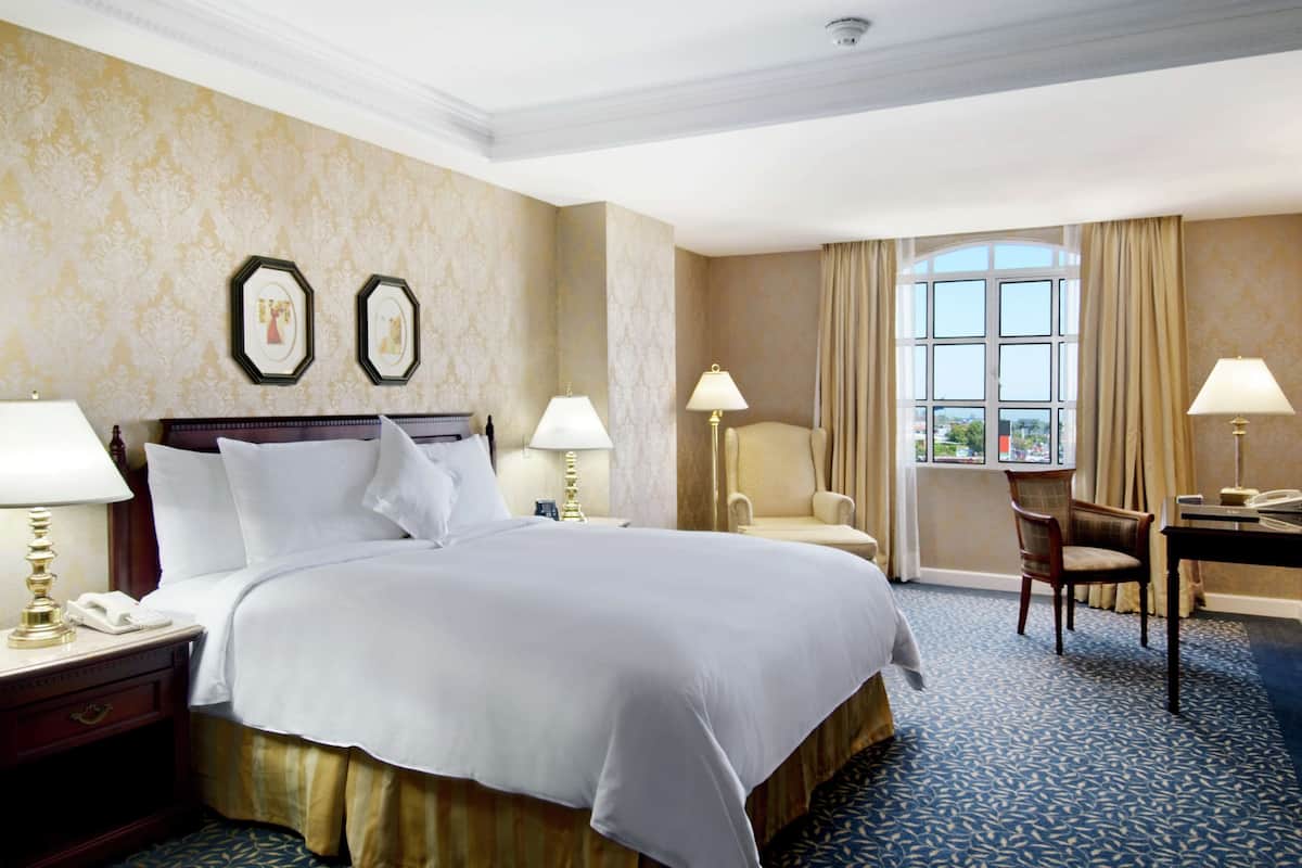 A hotel room with a large bed, two bedside tables with lamps, a desk with a chair, and a large window with curtains.