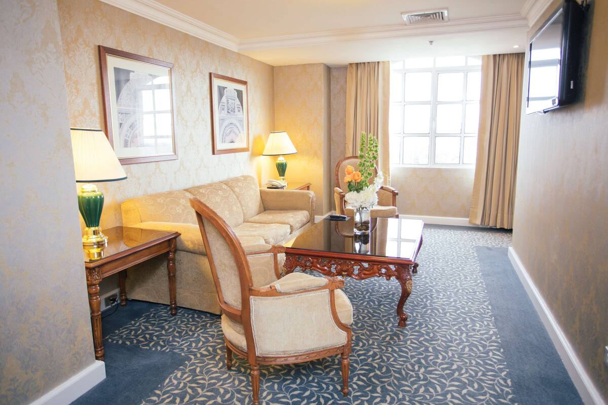 A hotel room with a sofa, armchair, coffee table, lamp, and a large window with curtains.