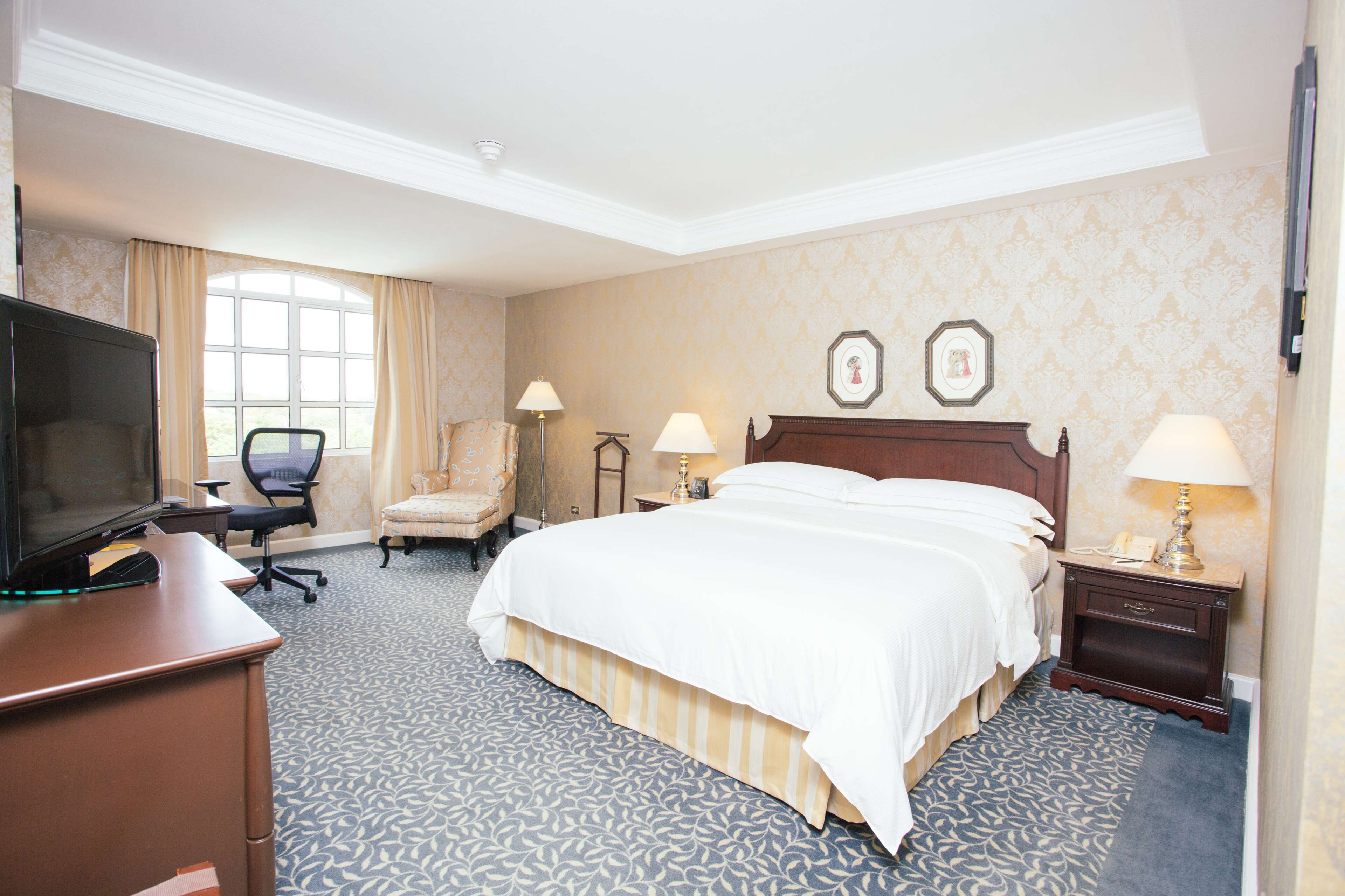 A hotel room with a large bed, a desk with a TV, two bedside lamps, a chair, and a window with curtains.
