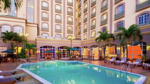 A hotel pool area with lounge chairs, palm trees, and a large building with balconies. - Hilton Princess Managua (Managua)