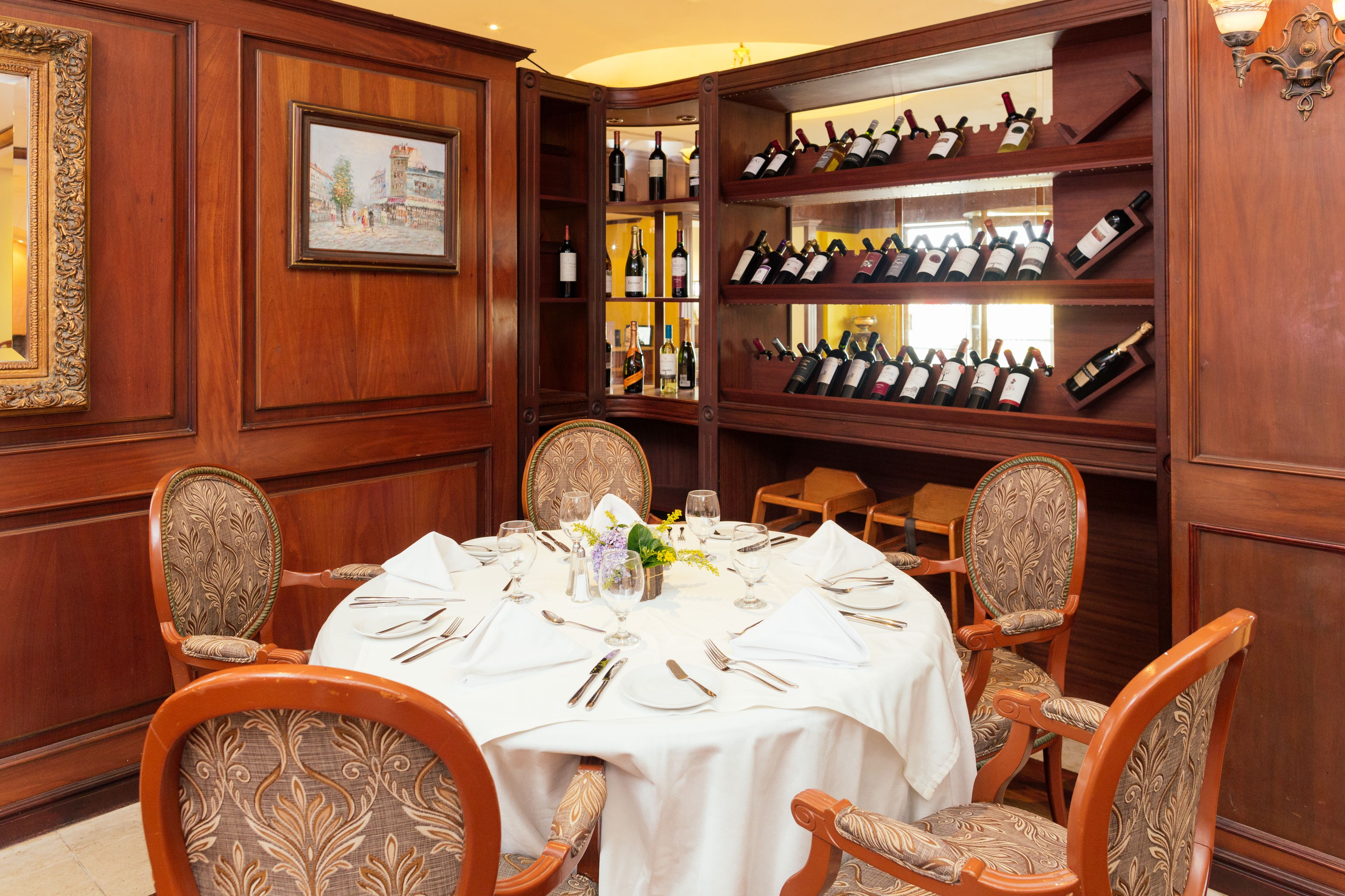A dining room with a round table set for six, surrounded by ornate chairs. A wine rack with multiple bottles is visible in the background.