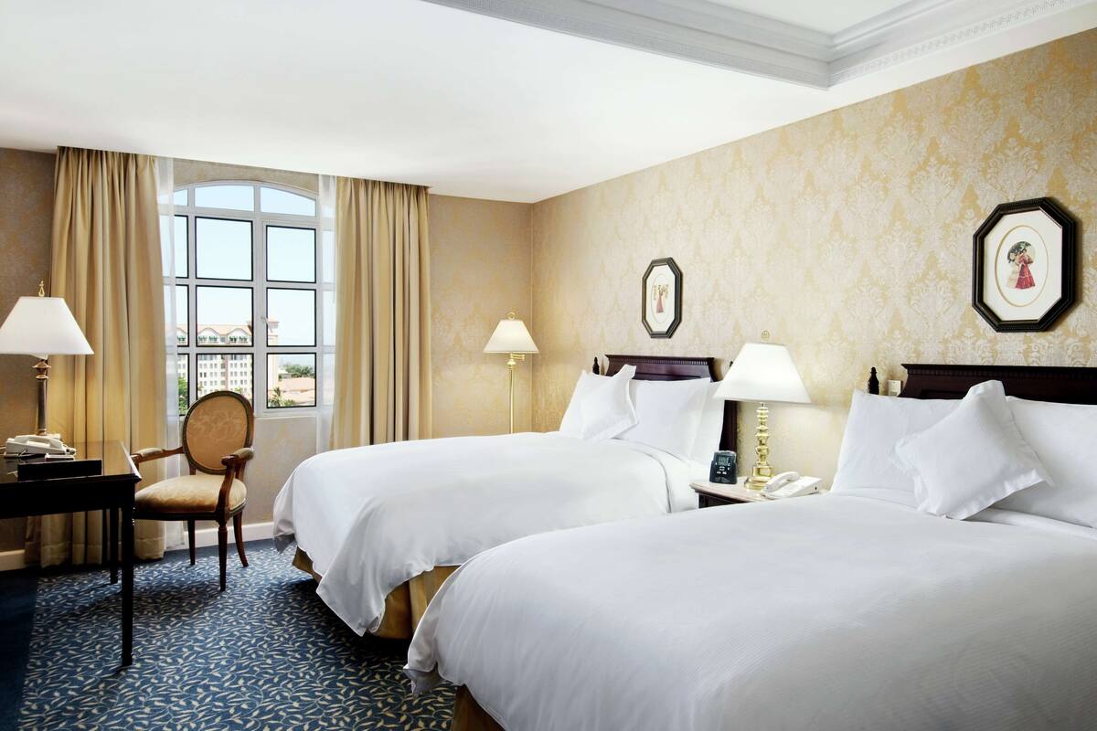 A hotel room with two beds, a desk, a chair, and a large window with curtains.