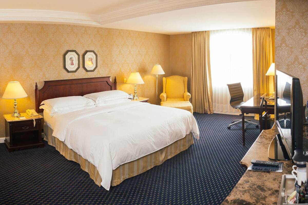 A hotel room with a large bed, two bedside lamps, a chair, a desk with a computer, and a television.