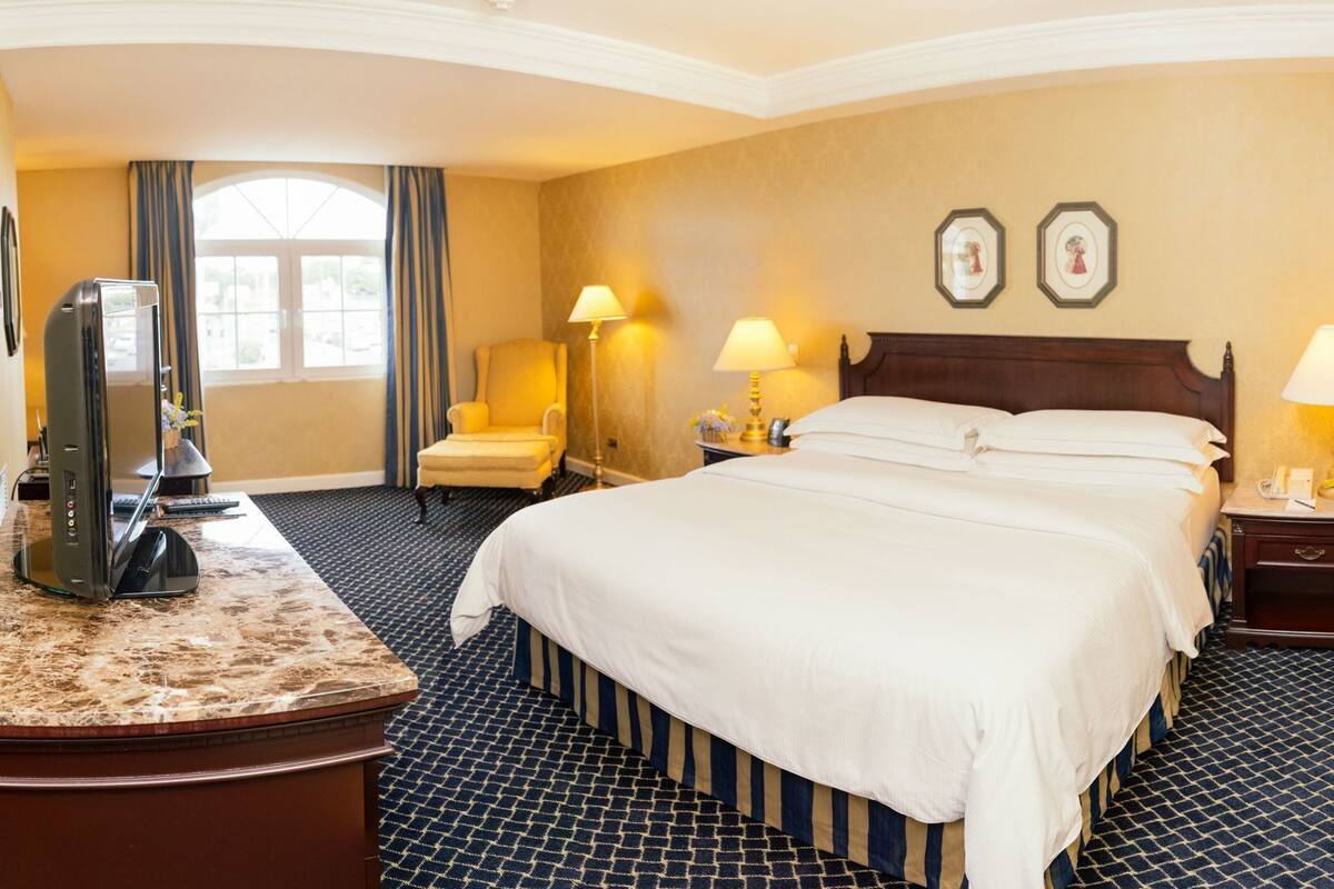 A hotel room with a large bed, a desk with a computer, two bedside lamps, a chair, and a window with curtains.