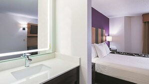 A hotel room with a large mirror, a white sink, a wooden vanity, a bed with white bedding, a bedside table with a lamp, and a wall-mounted TV. - La Quinta Inn & Suites by Wyndham Minneapolis Northwest (Brooklyn Park)
