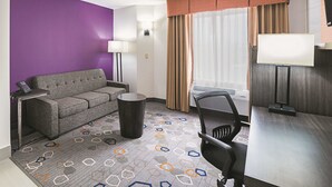 A hotel room with a purple wall, a grey sofa, a small round table, a black office chair, a lamp, and a window with curtains. - La Quinta Inn & Suites by Wyndham Minneapolis Northwest (Brooklyn Park)