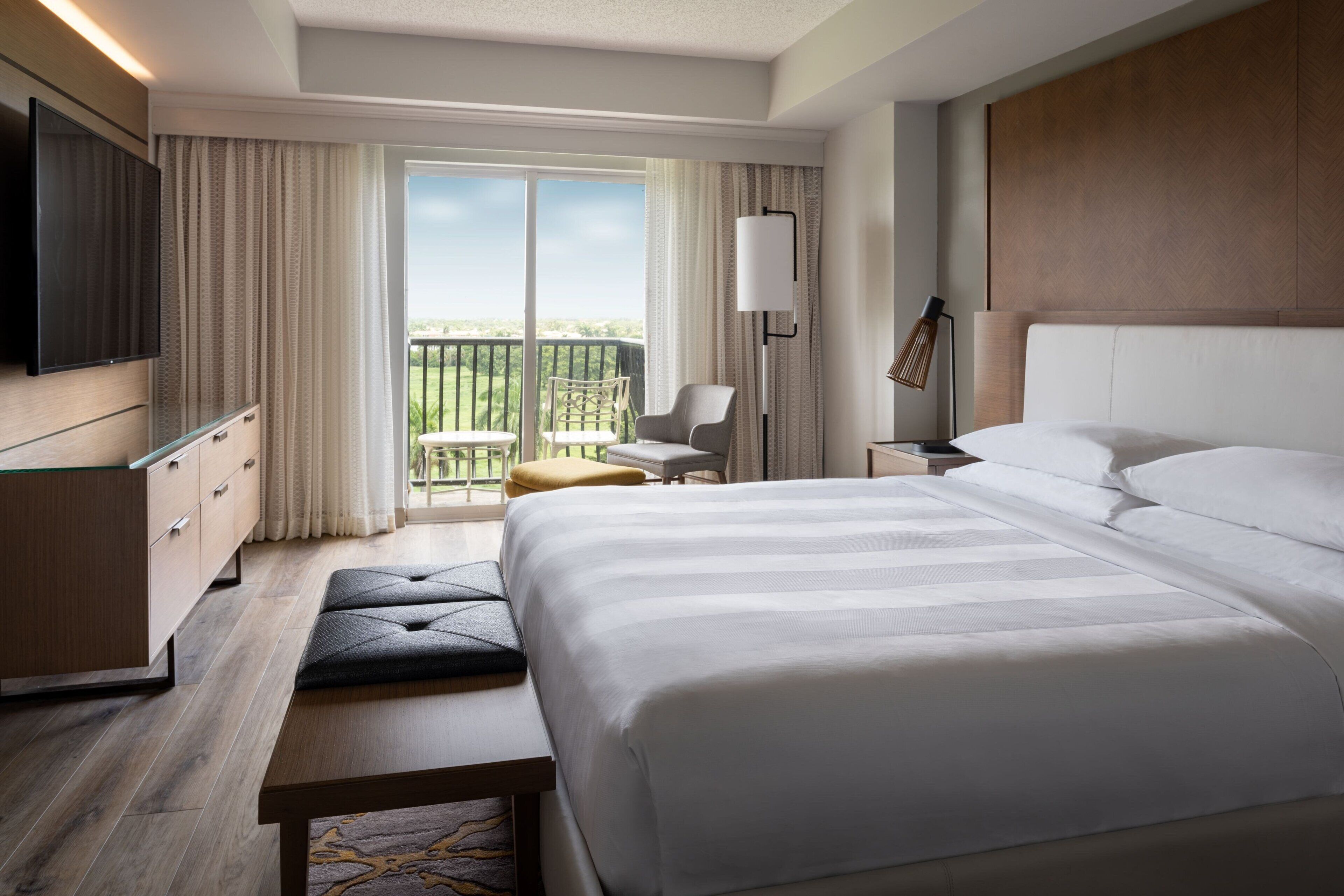 A hotel room with a large bed, a TV, a desk, a chair, and a balcony with a view.
