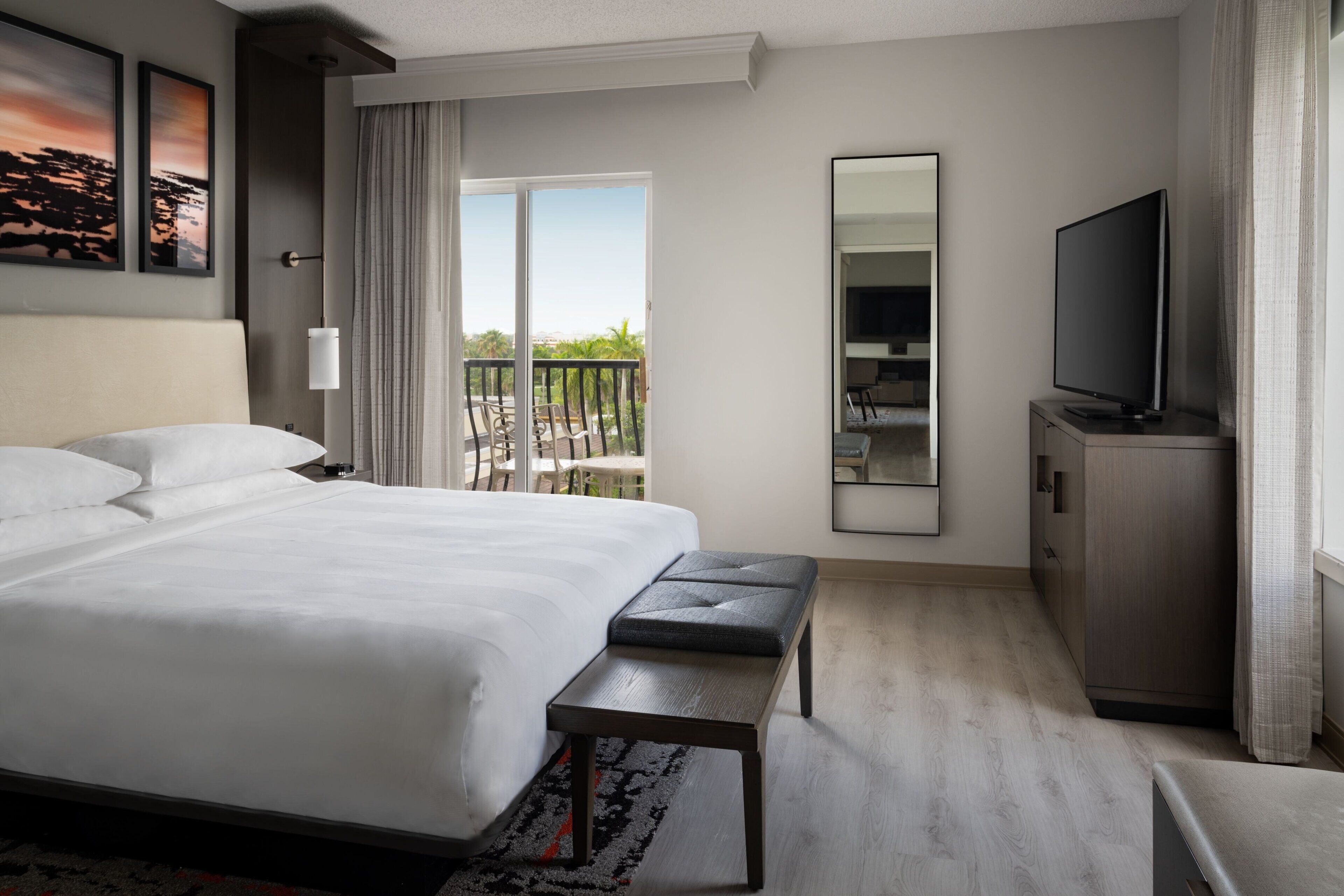 A hotel room with a large bed, a bench, a TV, a mirror, and a balcony with outdoor furniture.