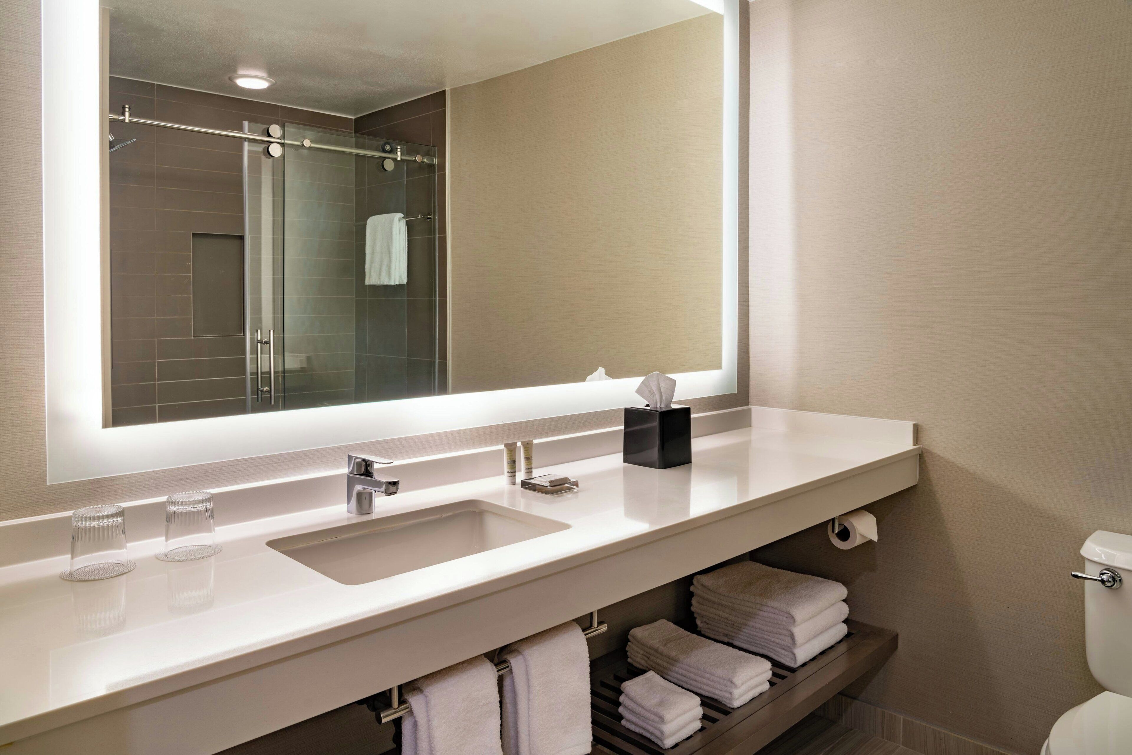 A modern bathroom with a large mirror, two sinks, a shower, and a toilet.