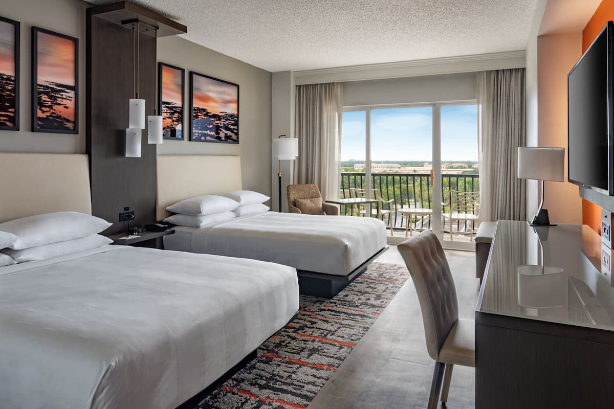 A hotel room with two beds, a desk, a TV, and a balcony with a view.
