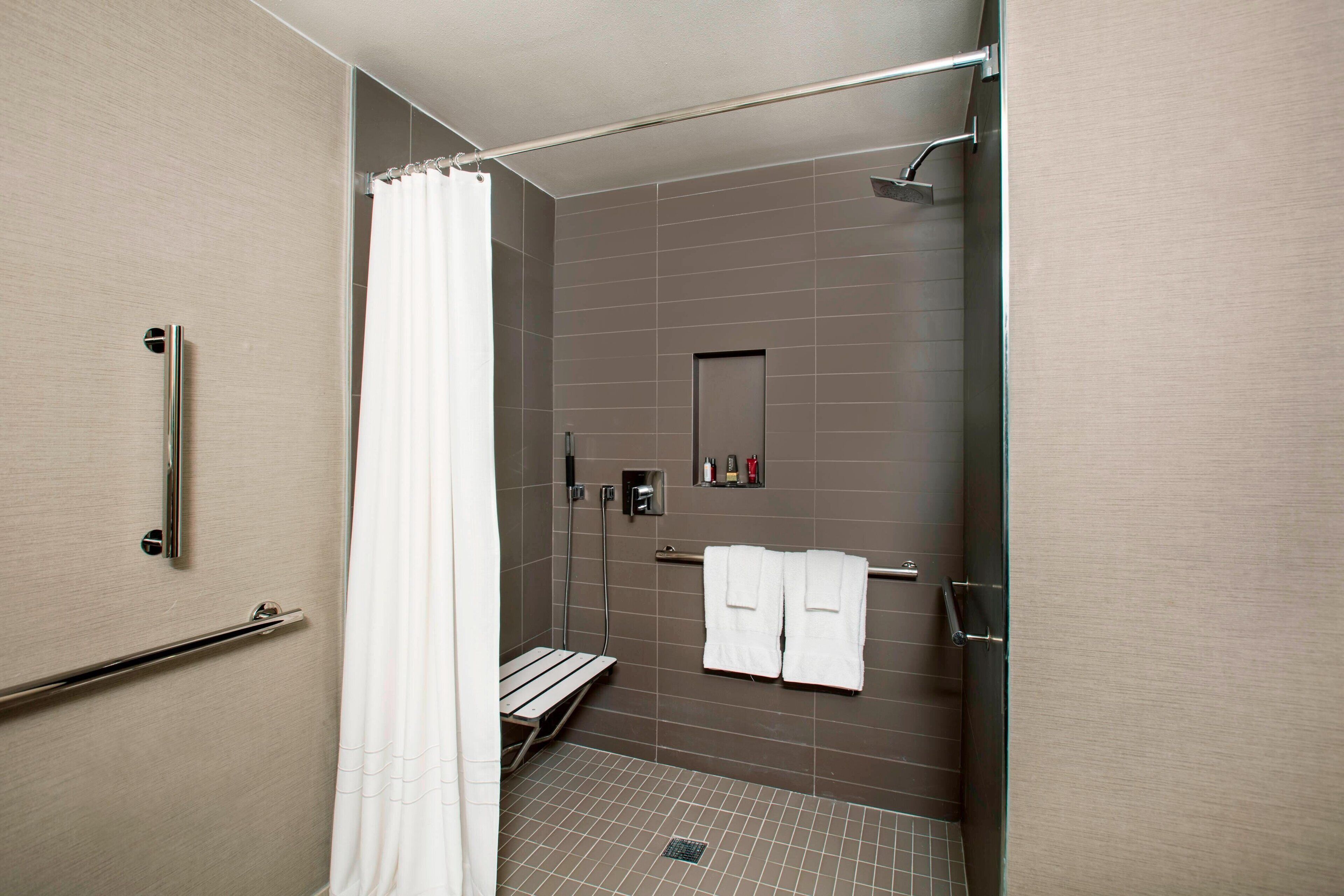 Un espace douche moderne doté d’une porte en verre, d’un rideau de douche blanc, d’un banc et d’un porte-serviettes avec deux serviettes.