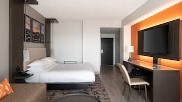 A modern hotel room with a bed, a desk with a chair, a television, and artwork on the wall.