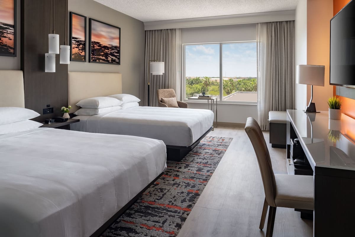 A hotel room with two beds, a desk with a chair, a TV, and a window with a view of the outdoors.