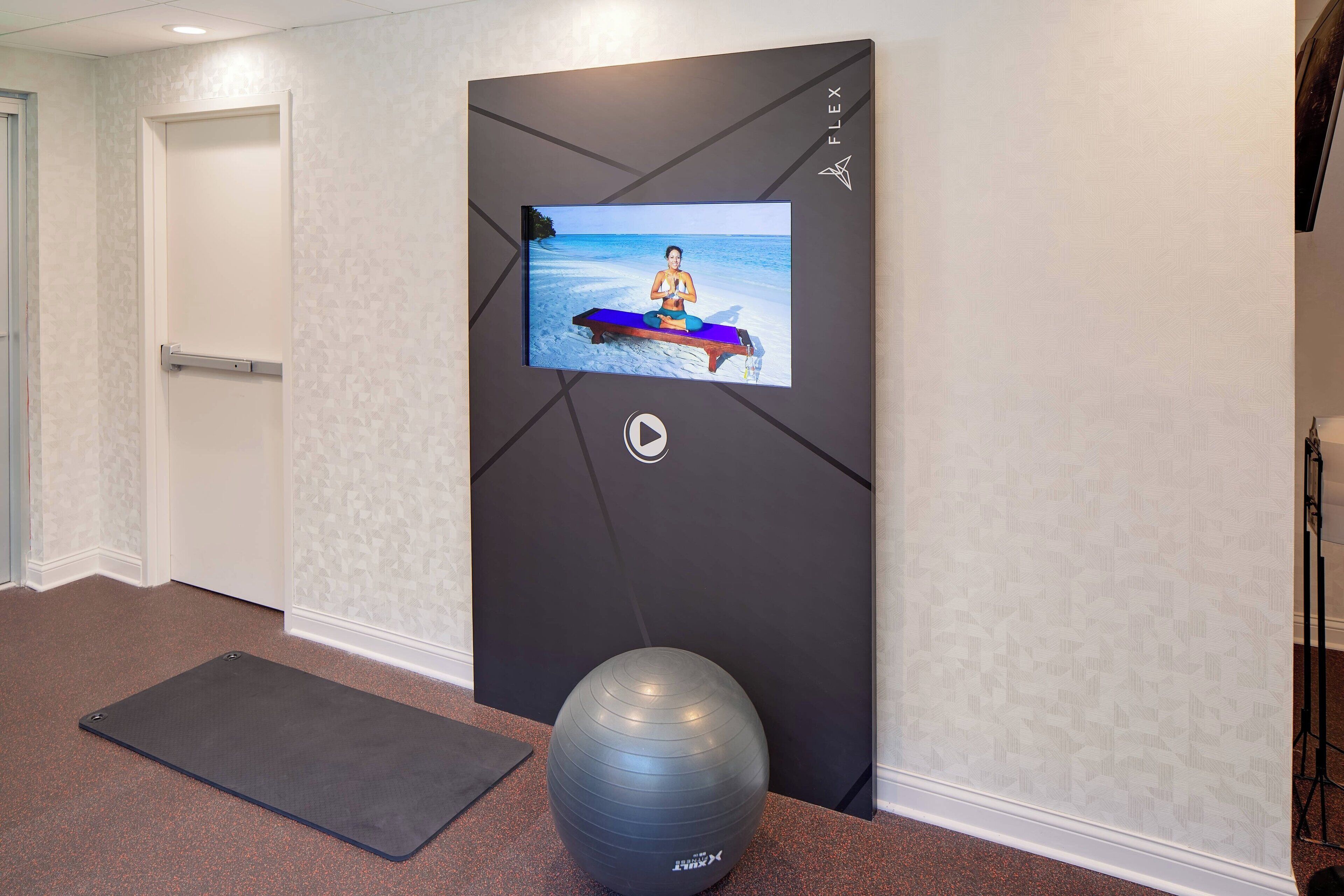 A hallway with a yoga mat, a stationary exercise ball, and a TV screen displaying a yoga pose.