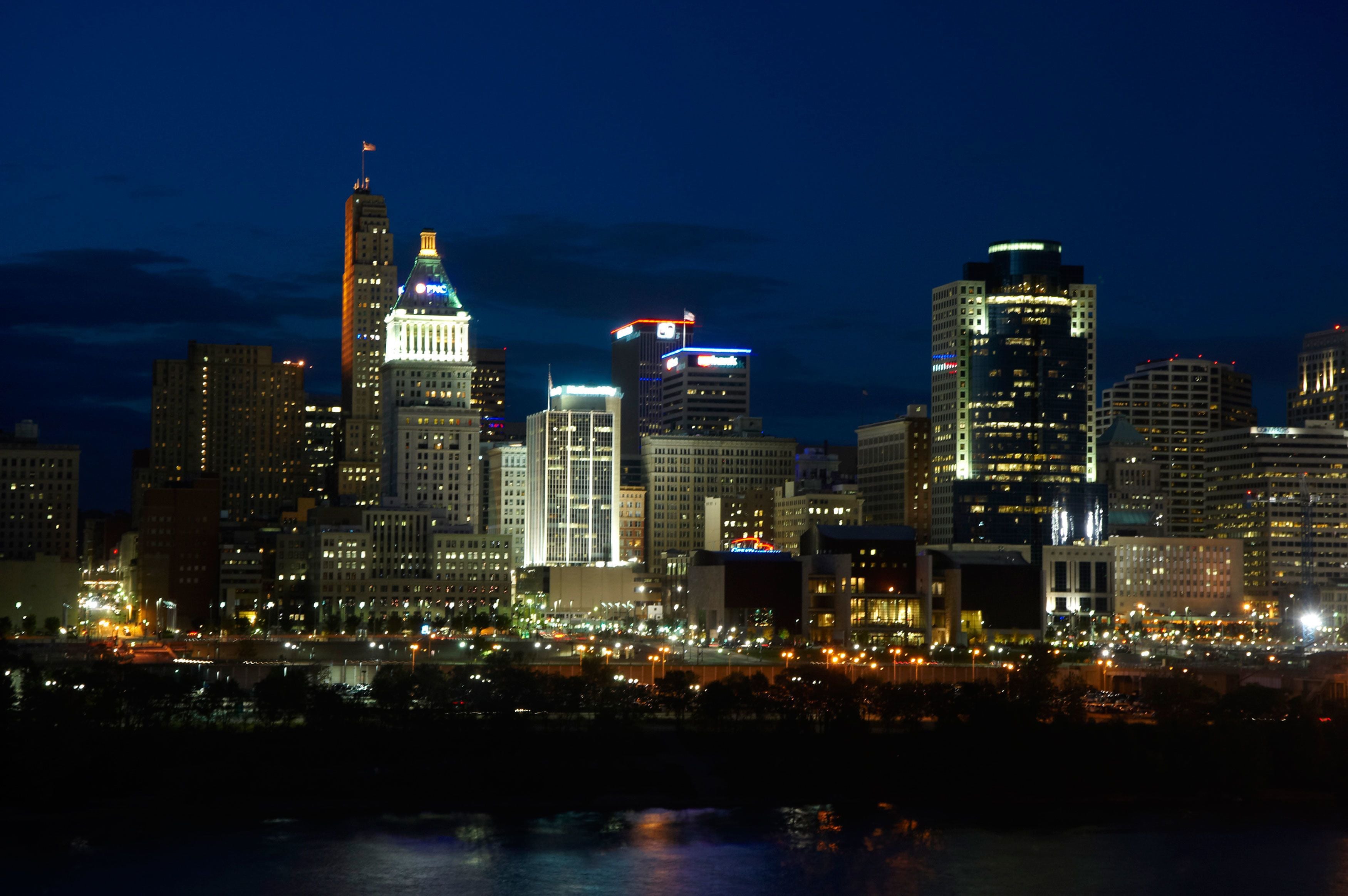 A nighttime city skyline with illuminated buildings and a river in the foreground.