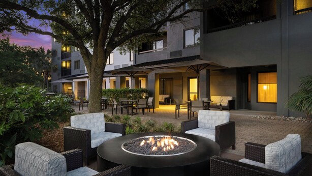 A courtyard with a fire pit surrounded by wicker furniture, a tree, and a building with multiple balconies and windows. - Courtyard by Marriott Myrtle Beach Barefoot Landing (Myrtle Beach)