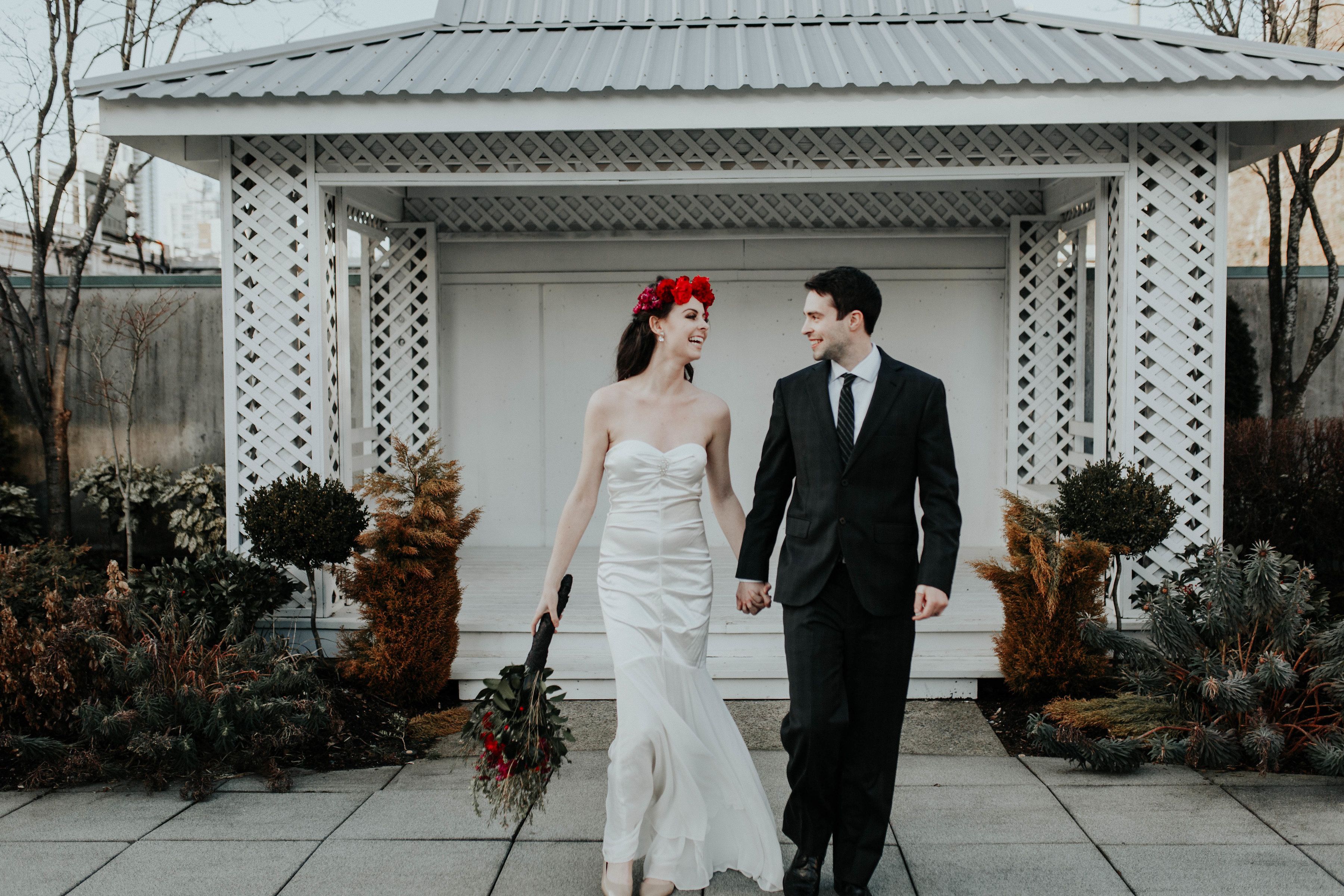 A couple walking hand in hand in front of a white gazebo with a lattice design, surrounded by evergreen shrubs.