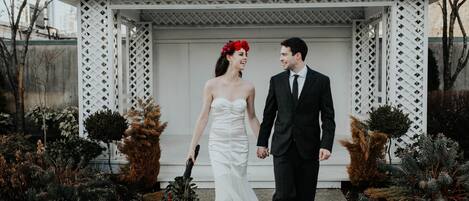 A couple walking hand in hand in front of a white gazebo with a lattice design, surrounded by evergreen shrubs.