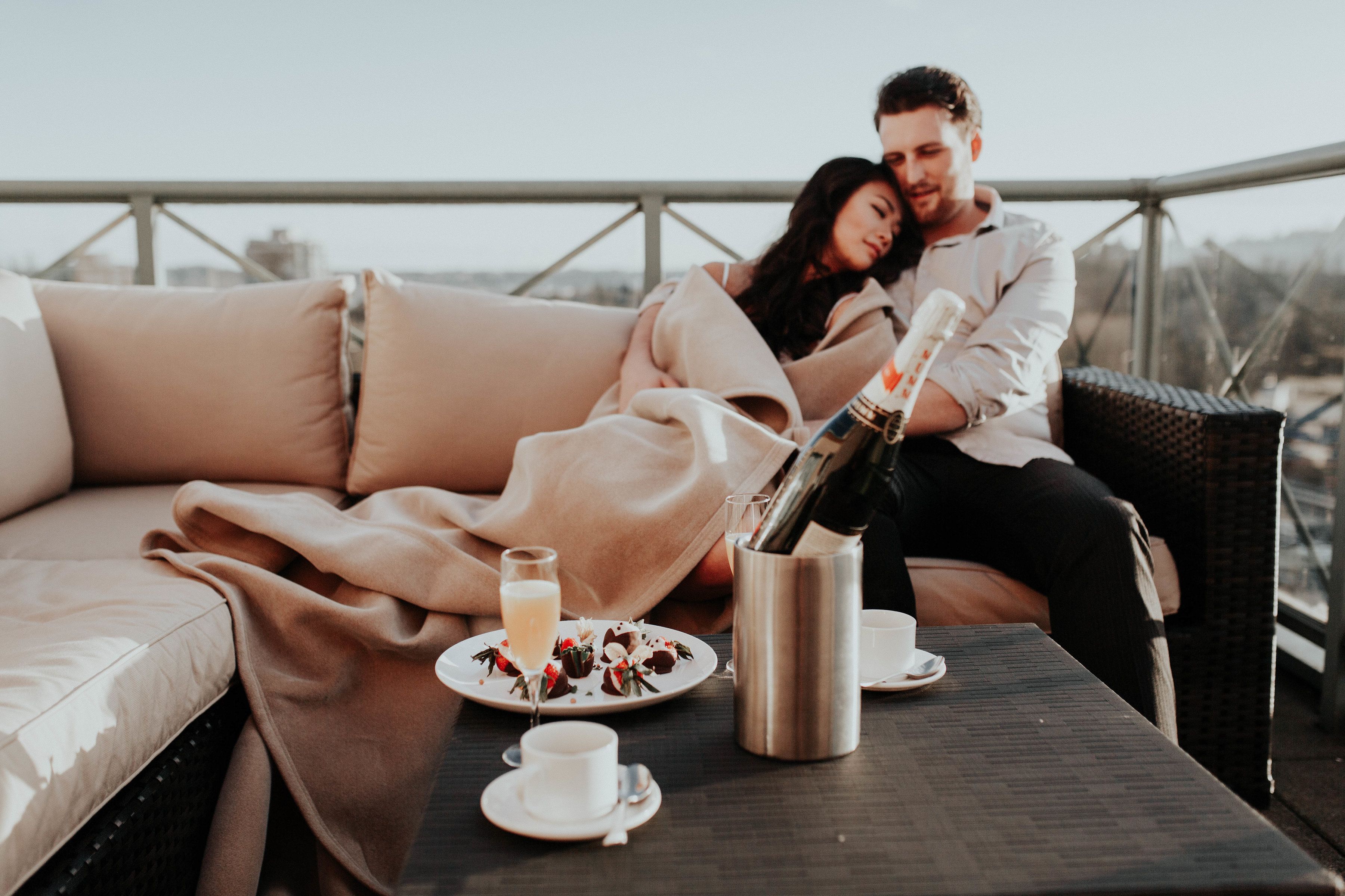 A couple sitting on a cushioned outdoor sofa with a bottle of champagne, a glass of wine, and a plate of food on a table.