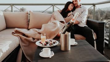 A couple sitting on a cushioned outdoor sofa with a bottle of champagne, a glass of wine, and a plate of food on a table.