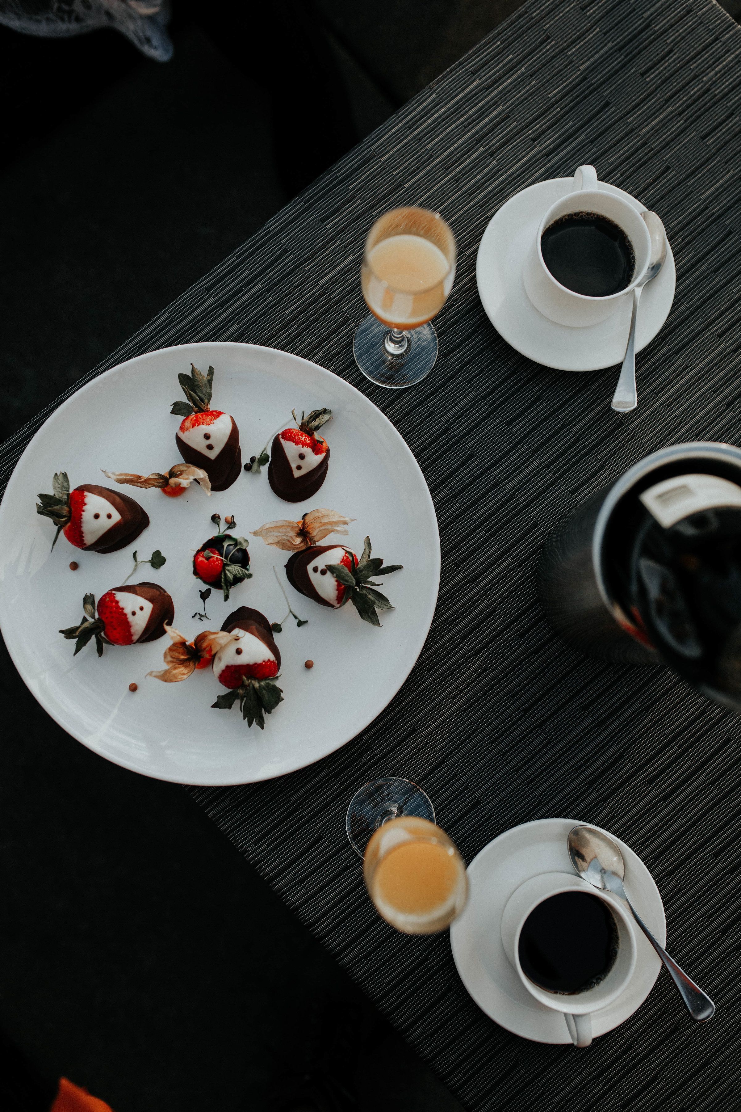 Una mesa con un plato de fresas cubiertas de chocolate, una taza de café y un vaso de jugo de naranja.