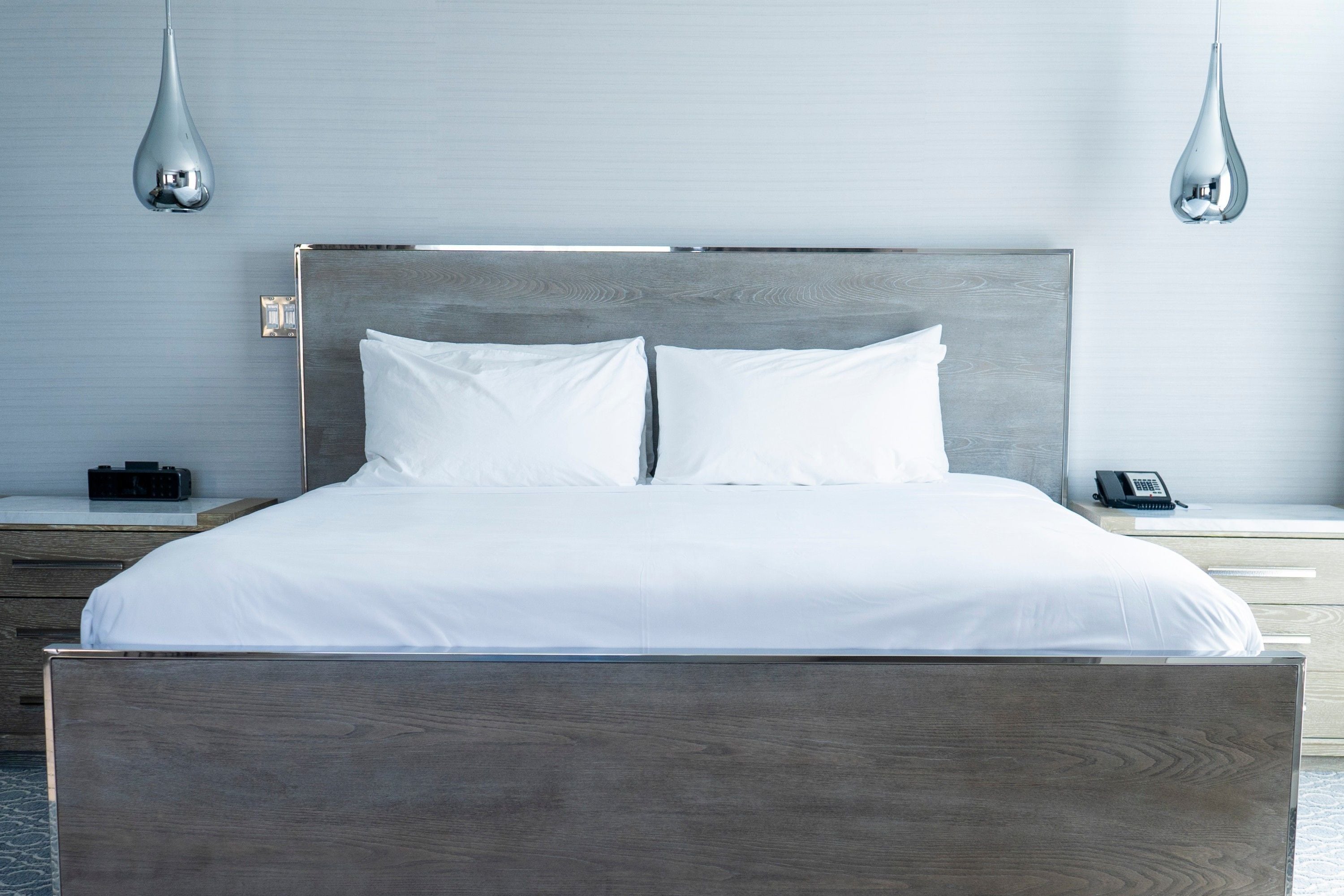 A modern hotel room with a wooden bed frame, white bedding, and a bedside table with a phone and a small black object.