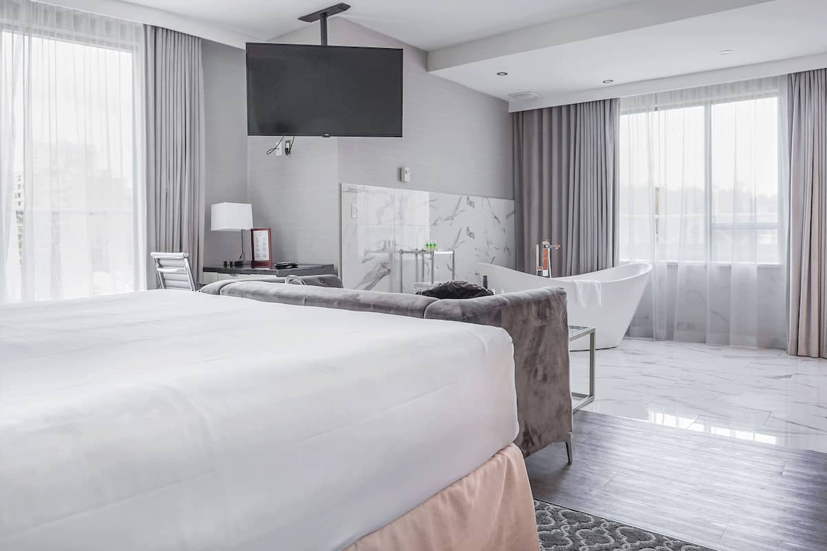 Ett modernt hotellrum med en stor säng, en sittgrupp med en fåtölj, en platt-TV som är monterad på väggen, en vägg beklädd med marmorplattor och ett stort fönster med gardiner.