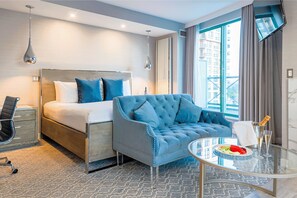 A hotel room with a bed, a blue armchair, a glass table with a plate of fruit and champagne, and a city view through large windows.