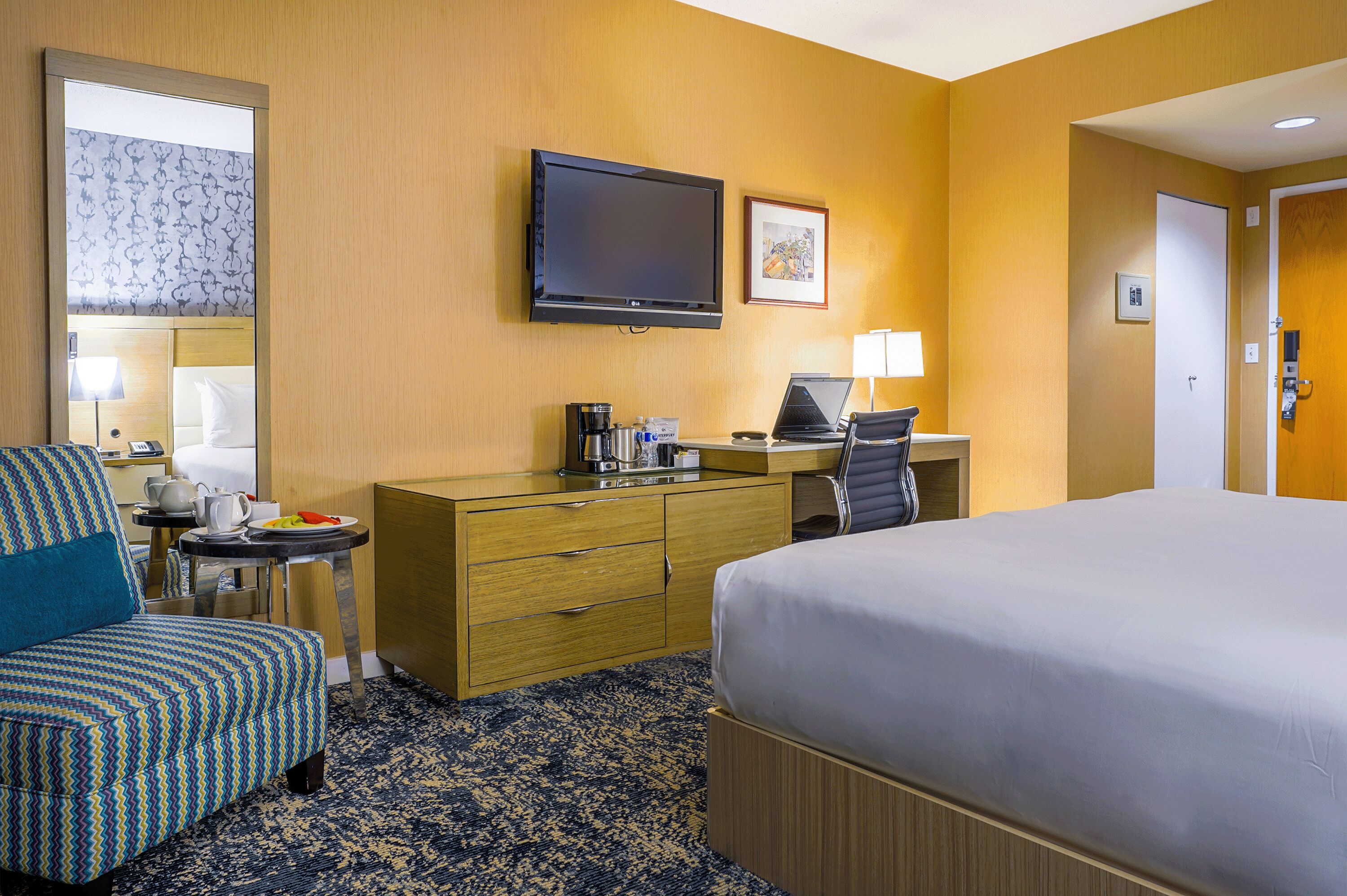 A hotel room with a bed, a desk with a computer, a chair, a coffee maker, and a TV on the wall.