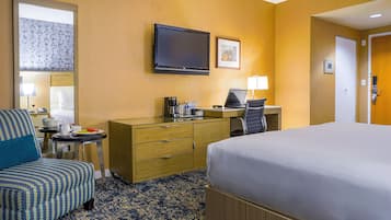 A hotel room with a bed, a desk with a computer, a chair, a coffee maker, and a TV on the wall.